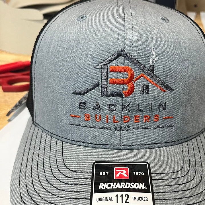 Embroidered home builder logo on a heather grey and black Richardson 112 trucker hat