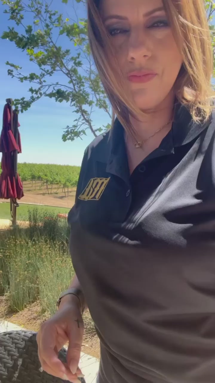 Woman outdoors wearing a black polo with the gold MyCali Designs logo embroidered on the chest